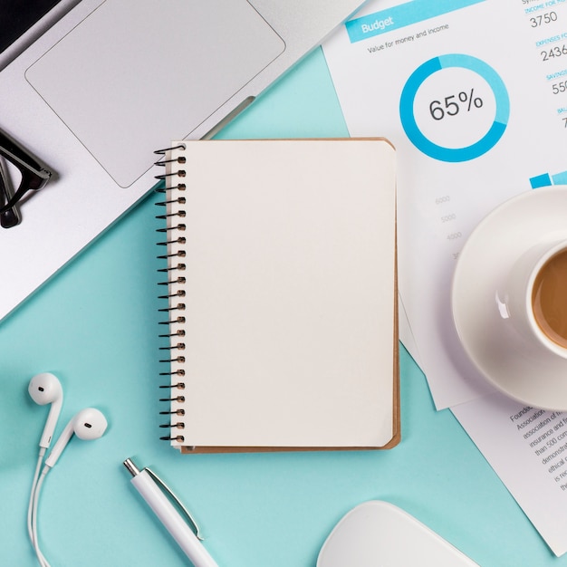 Bloco de notas em espiral fechado com laptop, fone de ouvido, caneta, mouse e xícara de café no plano de orçamento na mesa azul