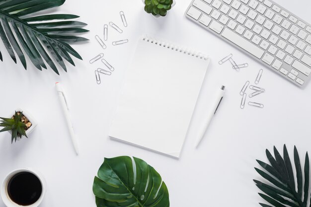 Bloco de notas espiral em branco com o teclado; clipes de papel e folhas verdes sobre fundo branco
