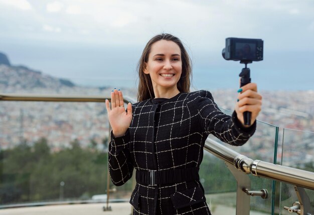 Blogueira sorridente está tirando selfie mostrando oi gesto para sua mini câmera no fundo da vista da cidade