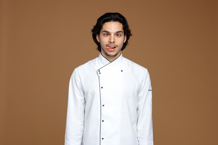 Foto grátis bobo jovem chef masculino vestindo uniforme olhando para a câmera com os olhos cruzados, mostrando a língua isolada no fundo marrom