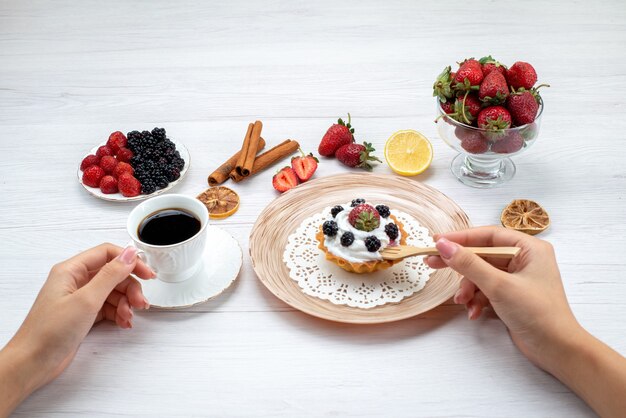 Bolo cremoso delicioso com frutas vermelhas sendo comido por uma mulher com café com canela em uma mesa clara, foto colorida