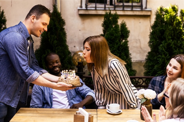 Bolo de aniversário com velas acesas que garota está soprando e melhores amigas no terraço de um café acolhedor