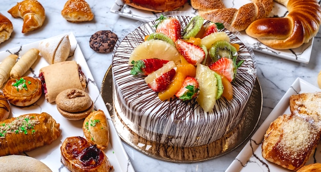 Bolo de frutas de vista lateral com creme de baunilha chocolate kiwi laranja morango abacaxi e bolos em cima da mesa