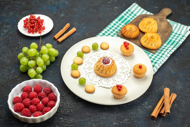 Bolo pequeno com biscoitos cremosos e uvas verdes, framboesas e cranberries na mesa escura