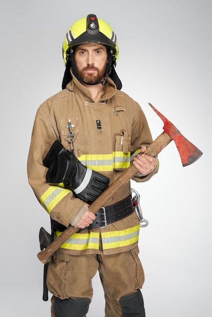 Bombeiro caucasiano barbudo com equipamento completo segurando machado vermelho para combate a incêndio na vista frontal do estúdio