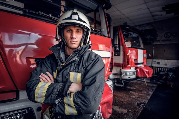 Foto grátis bombeiro confiante vestindo uniforme de proteção ao lado de um carro de bombeiros em uma garagem de um corpo de bombeiros, braços cruzados e olhando para uma câmera