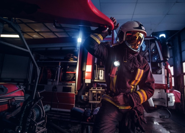 Bombeiro vestindo uniforme de proteção ao lado de um carro de bombeiros em uma garagem de um corpo de bombeiros