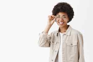 Foto grátis bonita, feliz e feminina afro-americana com penteado afro, tocando a armação dos óculos e sorrindo, confiante de que pode fazer qualquer coisa