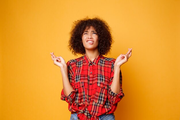 Bonita fêmea negra emocional cruza os dedos, espera que todos os desejos se tornem realidade sobre fundo laranja brilhante. Pessoas, linguagem corporal e felicidade.