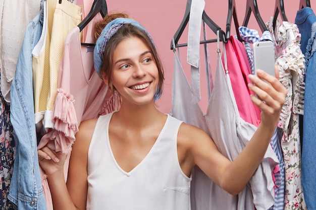 Bonita mulher sorridente tendo auto-retrato no celular genérico, posando em seu guarda-roupa, vangloriando-se sobre novos tops elegantes e vestidos que ela comprou na venda esta manhã