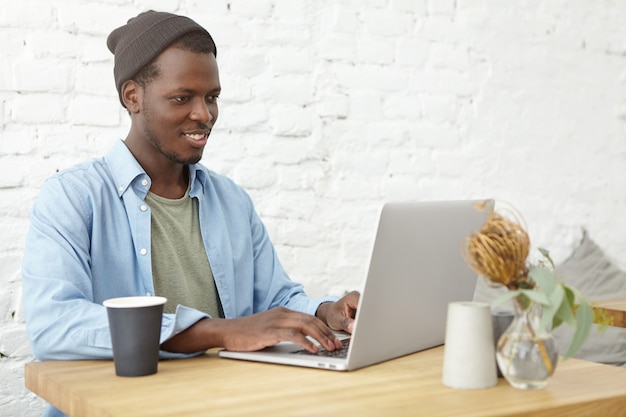 Bonitão afro-americano sentado no refeitório na frente do laptop aberto, digitando e pesquisando na internet, bebendo café. Estudante do sexo masculino jovem de pele escura, preparando-se para as aulas na cafeteria