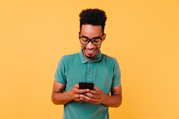 Bonitão negro de óculos grandes, lendo a mensagem do telefone. Retrato de homem Africano satisfeito segurando o smartphone.