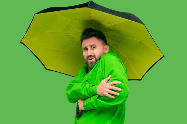 Bonito barbudo congelamento jovem segurando guarda-chuva e olhando para a câmera isolada no estúdio verde na moda vívido.