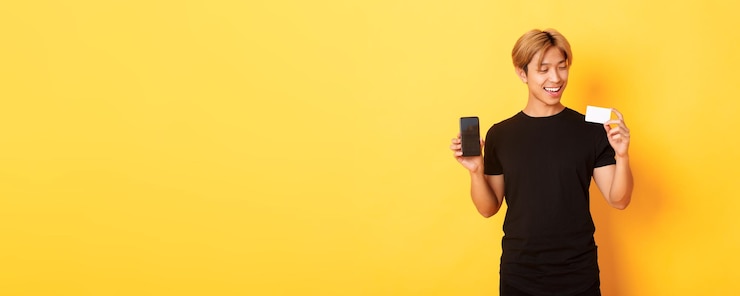 Foto grátis bonito cara asiático satisfeito segurando o cartão de crédito e mostrando a tela do smartphone sorrindo de pé satisfeito