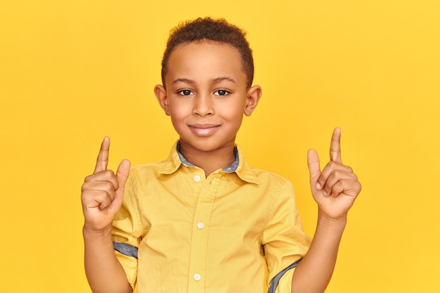 Foto grátis bonito e alegre garoto afro-americano com uma expressão facial feliz, sorrindo para a câmera, levantando os dois dedos indicadores, apontando para cima.