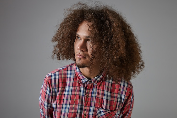 Foto grátis bonito e atraente jovem mestiço com a barba por fazer, pele bronzeada, cabelo cacheado solto e piercing no nariz posando, olhando para longe com expressão facial séria, vestido com camisa xadrez