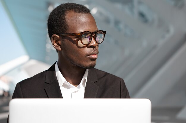 Bonito e bem sucedido jovem trabalhador corporativo afro-americano de óculos e terno preto, sentado ao ar livre na frente do laptop, olhando para longe, tendo expressão pensativa, absorvido em questões de negócios