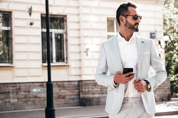 Bonito e confiante hipster elegante modelo lambersexual Homem moderno vestido de terno branco Sexy moda masculina posando na rua na cidade da Europa ao pôr do sol em óculos de sol olhando para smartphone