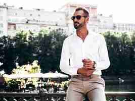 Foto grátis bonito e elegante hipster lambermodelo sexual homem moderno sexy vestido com camisa branca e calças moda masculina posando na rua perto do aterro na cidade do parque europa ao pôr do sol em óculos de sol