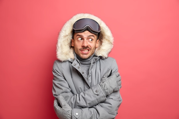 Foto grátis bonito e intrigado homem com a barba por fazer shievers e treme durante o dia gelado odeia o frio do inverno usa óculos de snowboard e jaqueta cinza.