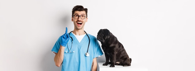 Foto grátis bonito e jovem médico na clínica veterinária apontando o dedo para cima e parecendo espantado em pé perto de um lindo p preto