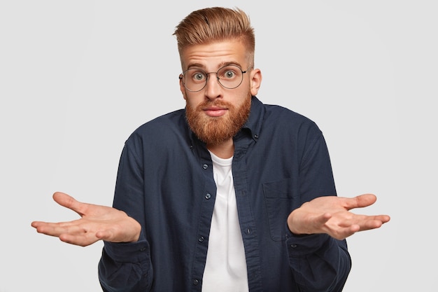 bonito e perplexo jovem ruivo com expressão sem noção, sente dúvida, escolhe entre duas coisas, usa óculos redondos, tem cabelo e barba de raposa, expressa hesitação