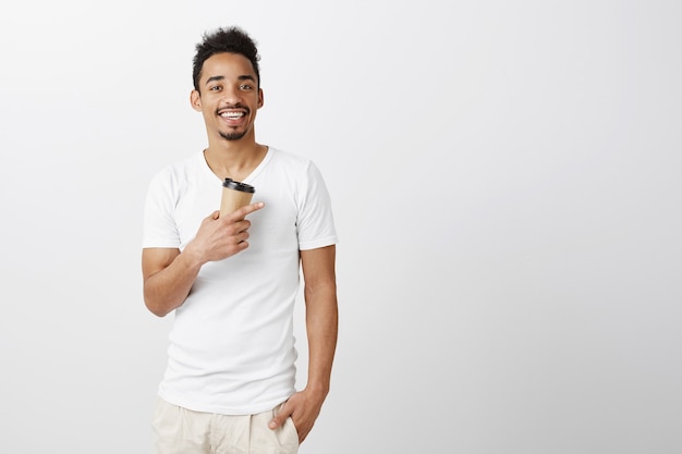 Bonito e sorridente homem afro-americano bebendo café e apontando para a direita, mostrando um café incrível