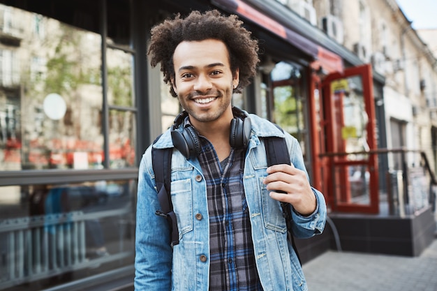 Bonito elegante afro-americano com penteado afro, vestindo casaco jeans e fones de ouvido, andando pela cidade.