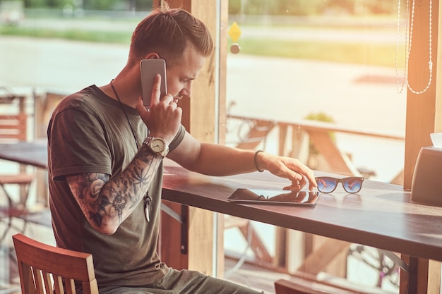 Bonito elegante hipster senta-se em uma mesa em um café na estrada, falando no smartphone.