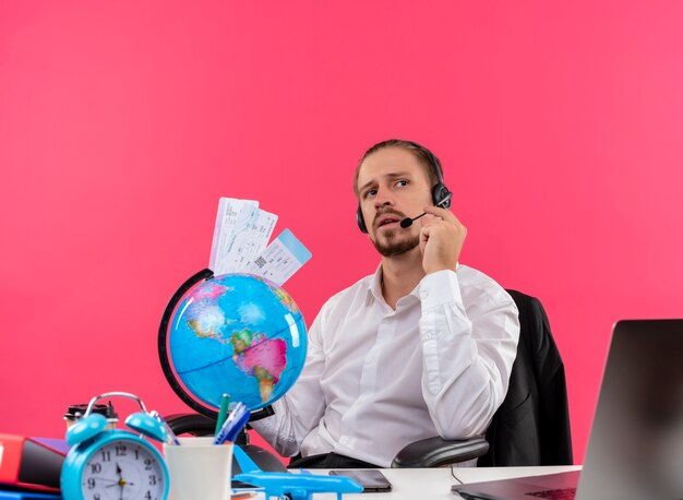 Bonito empresário de camisa branca e fones de ouvido com um microfone segurando passagens aéreas, ouvindo um cliente com cara séria sentado à mesa em escritório sobre fundo rosa