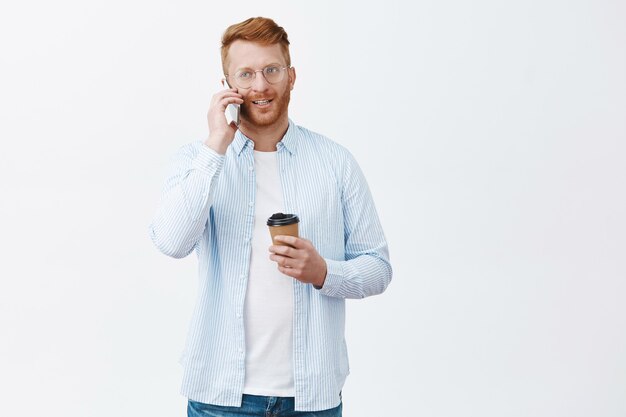 Bonito empresário moderno com cabelo ruivo de óculos, segurando um copo de bebida e falando casualmente por meio de uma parede cinza via smartphone