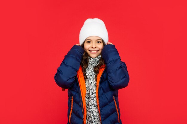 Bonito garoto positivo com cabelo encaracolado na jaqueta e chapéu. menina adolescente em fundo vermelho. retrato de criança vestindo roupas quentes com cachecol. expressar emoção positiva. moda de inverno.