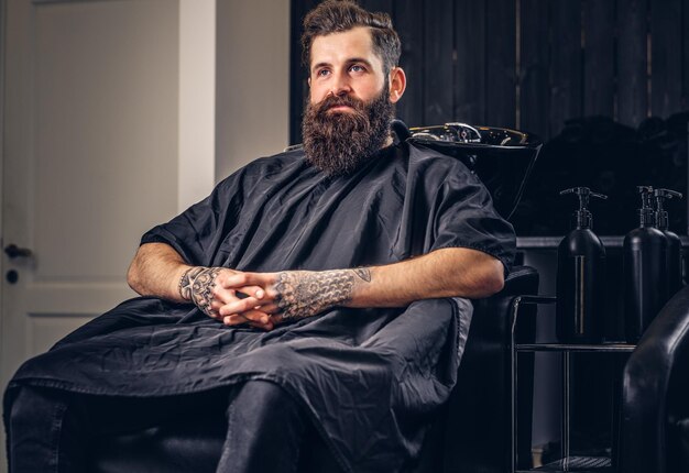 Bonito homem barbudo com uma tatuagem nos braços antes de lavar o cabelo em um salão de cabeleireiro.