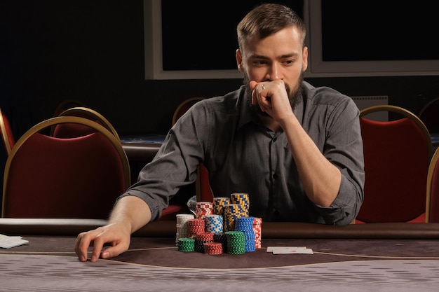 Foto grátis bonito homem barbudo de camisa cinza está jogando poker sentado à mesa no cassino. ele está fazendo apostas esperando por uma grande vitória. apostas por dinheiro. jogos de fortuna.