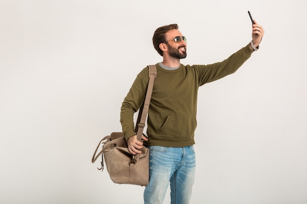 Bonito homem barbudo elegante em moletom com bolsa de viagem, jeans e óculos de sol isolados tirando foto de selfie no telefone