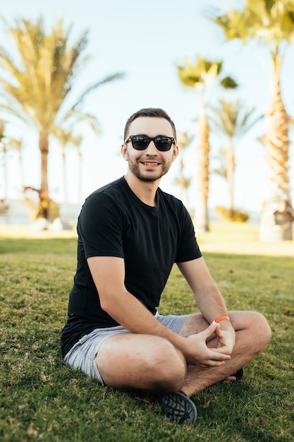 Bonito homem barbudo em óculos de sol, sentado na grama sob as palmas das mãos nas férias de verão.