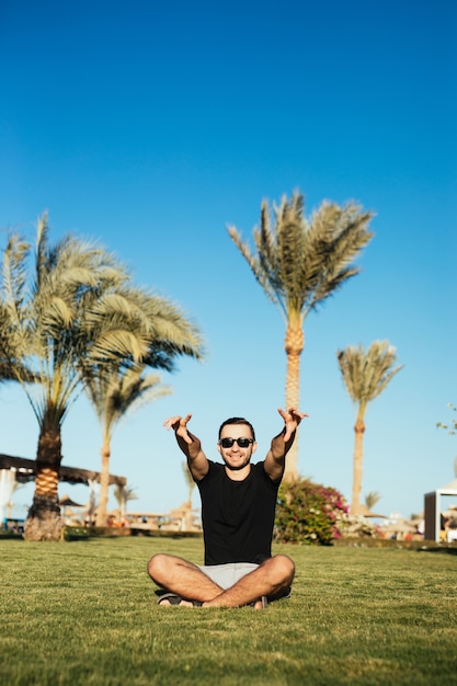 Bonito homem barbudo em óculos de sol, sentado na grama verde e relaxe, aproveite as férias de verão com as mãos levantadas, sorriso feliz.