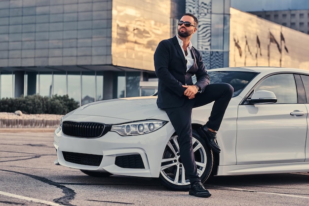 Bonito homem barbudo em óculos de sol, vestido com um terno preto, sentado em um carro de luxo contra um arranha-céu.