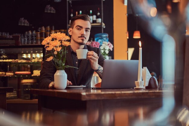 Bonito homem freelancer com barba elegante e cabelo vestido de terno preto, sentado em um café com um laptop aberto e segura uma xícara de café, olhando para uma câmera.