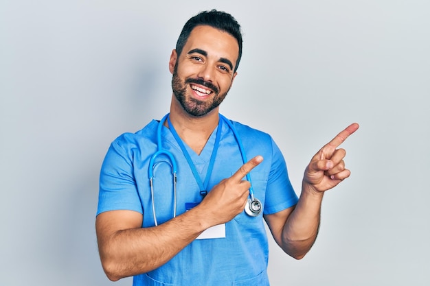 Bonito homem hispânico com barba vestindo uniforme médico sorrindo e olhando para a câmera apontando com as duas mãos e os dedos para o lado.