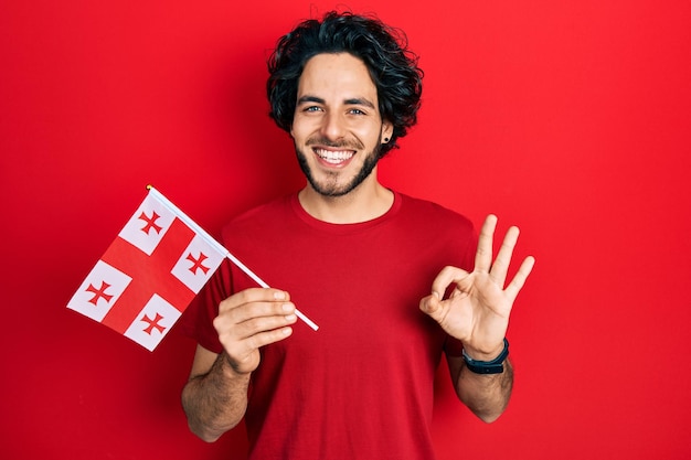 Bonito homem hispânico segurando a bandeira da geórgia fazendo sinal de ok com os dedos, sorrindo amigável gesticulando excelente símbolo