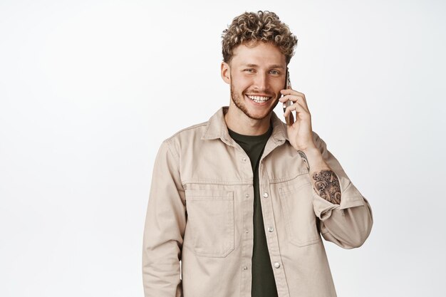 Bonito homem loiro sorridente falando no celular usando smartphone fazendo uma chamada em pé contra o fundo branco