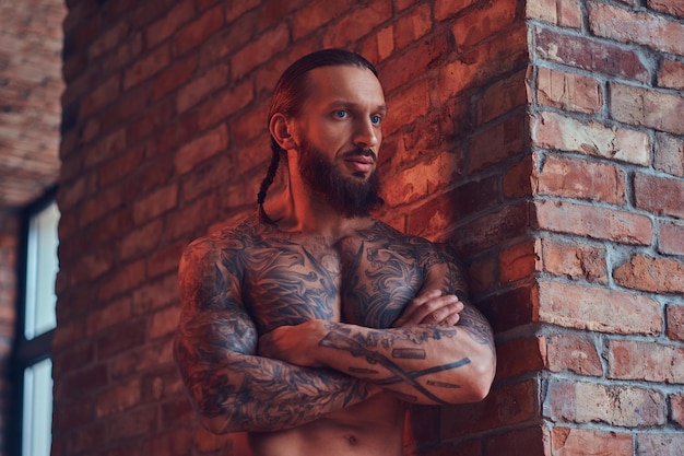 Bonito homem sem camisa tatuado com um corte de cabelo elegante e barba, de pé com os braços cruzados, encostado a uma parede de tijolos em uma sala com um interior loft.