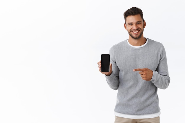 Bonito homem sorridente entusiasmado com cerdas no suéter cinza segurando a tela do smartphone de frente para a câmera apontando a tela do celular e sorrindo recomendo aplicativo carsharing ou site de compras