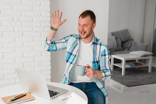 Bonito homem sorridente na camisa sentado na cozinha em casa na mesa trabalhando online no laptop