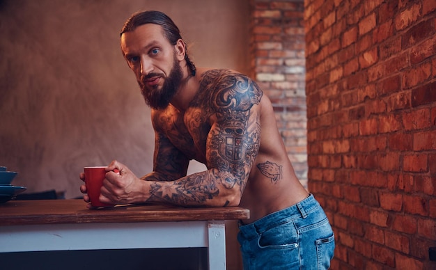 Foto grátis bonito homem tatuado barbudo com um corte de cabelo estiloso e corpo musculoso, bebe café, encostado em uma mesa.