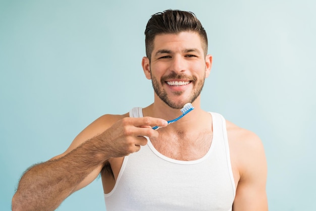 Bonito jovem macho escovando os dentes enquanto faz contato visual contra o fundo turquesa