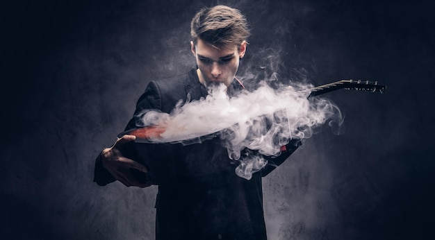 Bonito jovem músico com cabelo elegante em roupas elegantes exala fumaça em seu violão. Isolado em um fundo escuro.