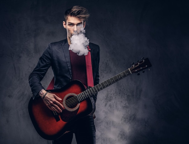 Bonito jovem músico com cabelo elegante em roupas elegantes exala fumaça enquanto toca violão. isolado em um fundo escuro.