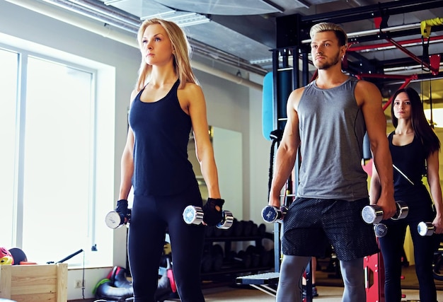 Foto grátis bonito loiro, homem atlético e dois modelos de fitness feminino magro fazendo exercícios de ombro com halteres em um clube de ginástica.